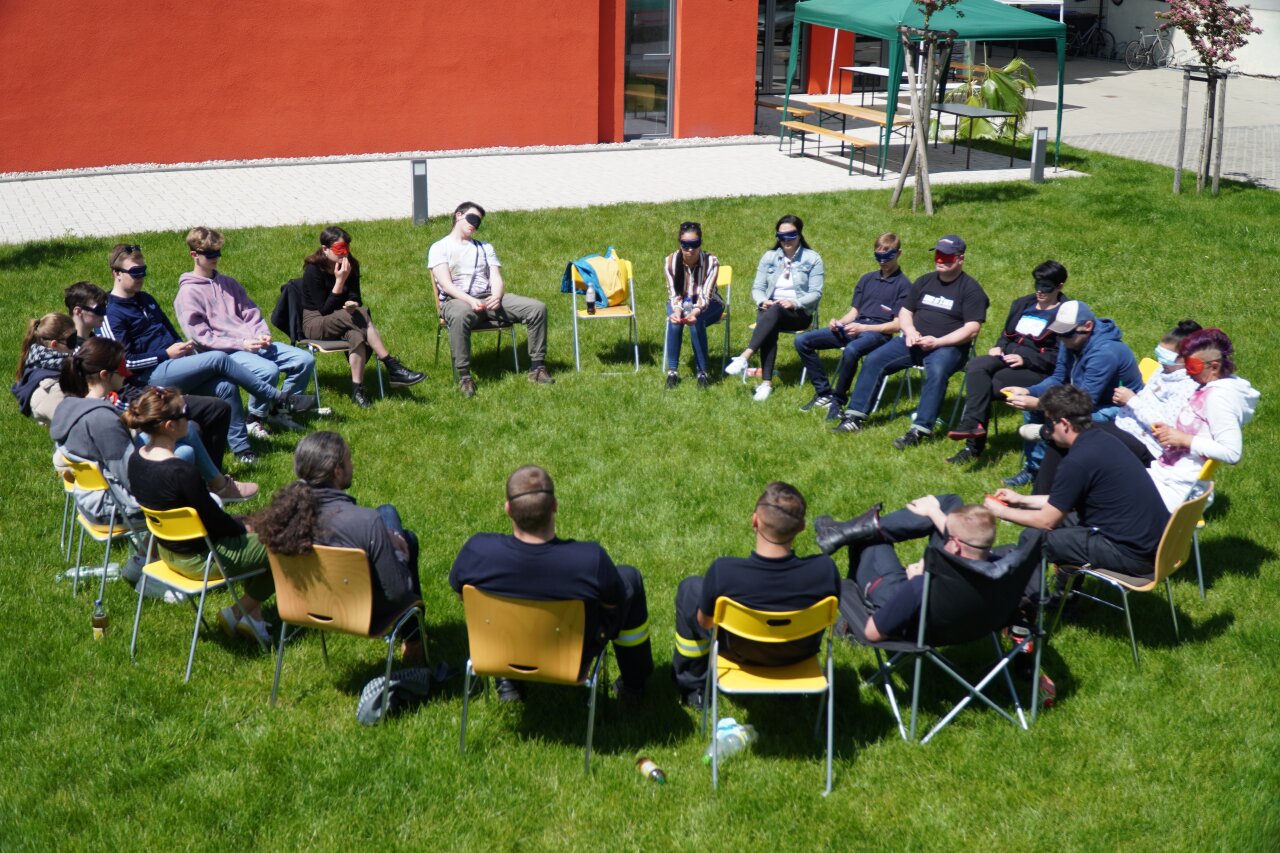 Viele Menschen sitzen mit verbundenen Augen im Stuhlkreis.